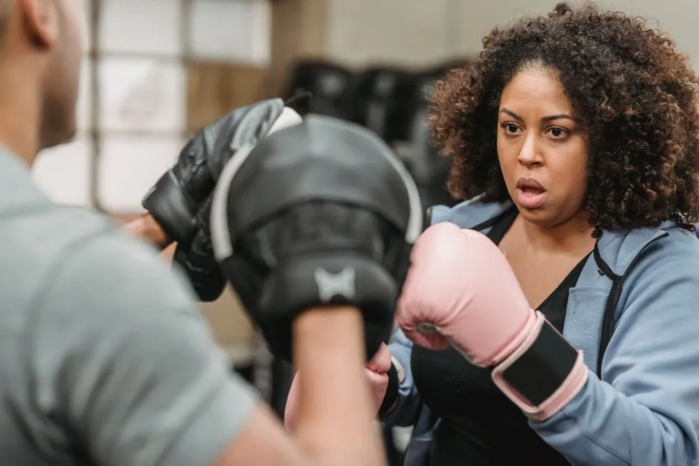 A woman in boxing gloves strikes pads held by a coach.