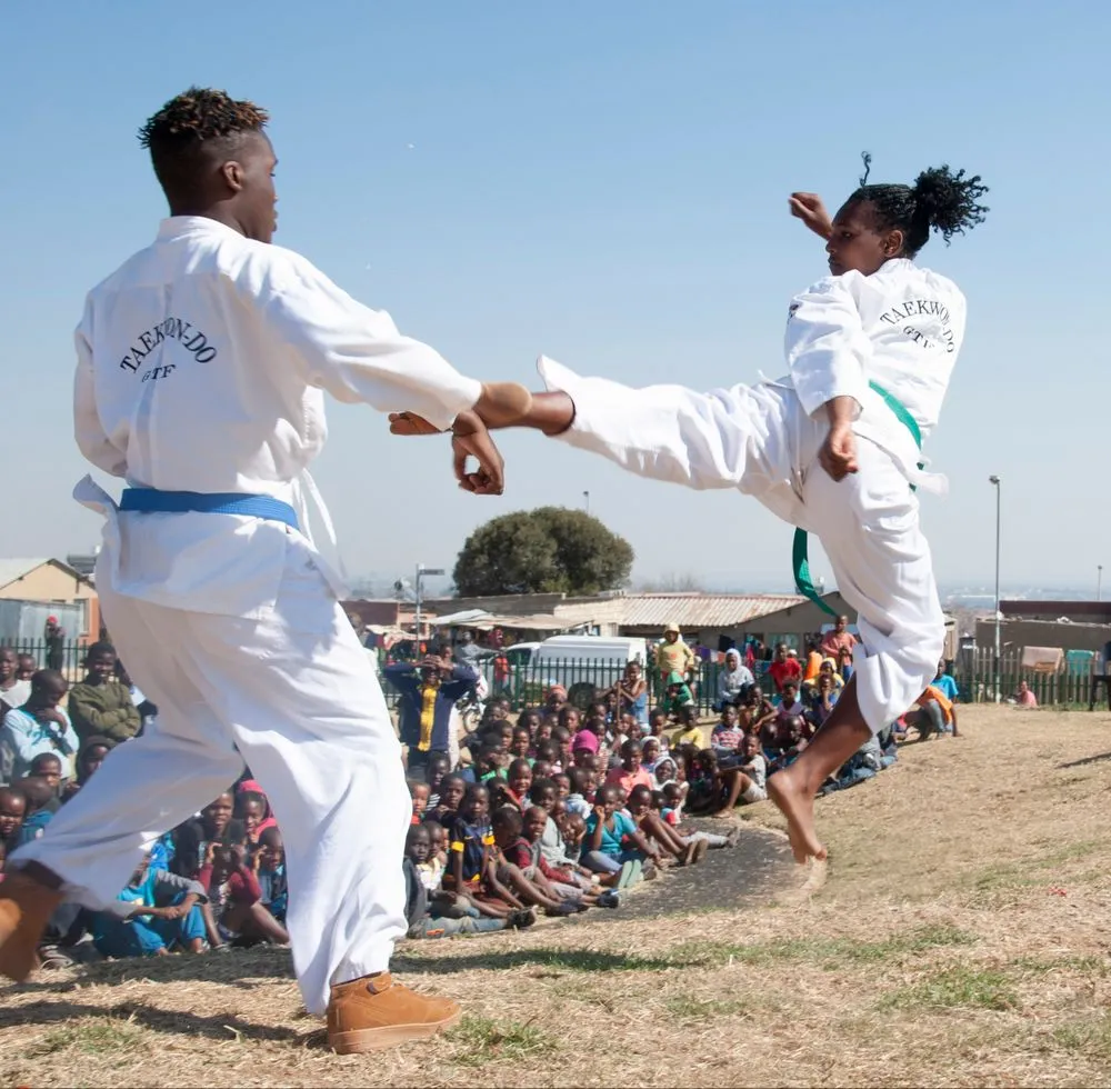 Two taekwondo athletes demonstrate a flying kick outdoors in front of a crowd.